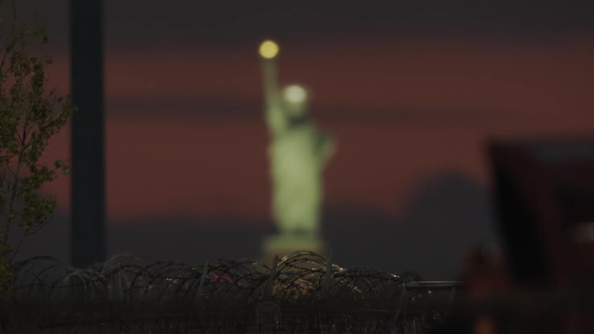 The statue of liberty is shining beyond a fence at dusk