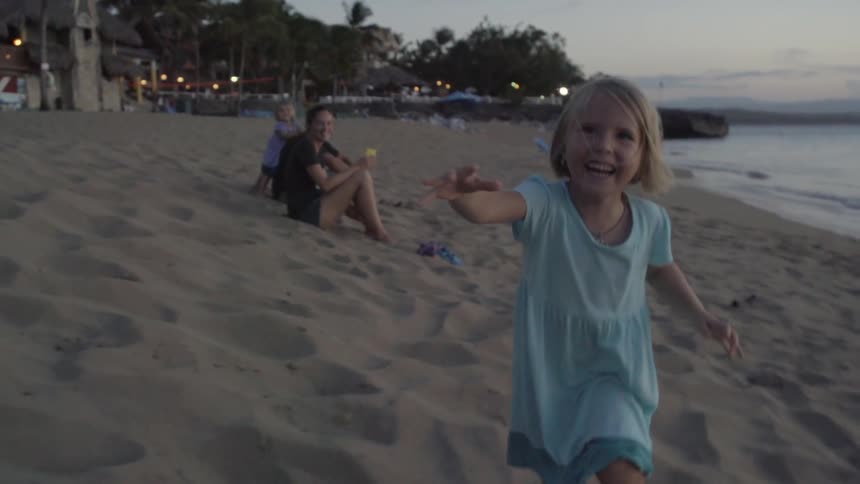 A laughing blonde girl in a blue dress runs down the beach