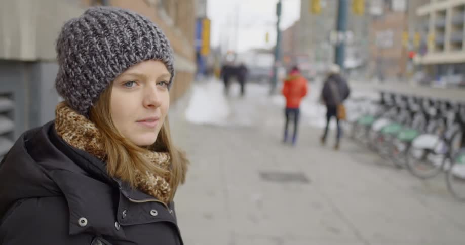 A woman stands on a city sidewalk with pedestrians walking in the background