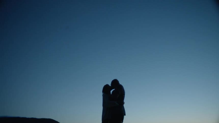A couple are kissing as they stand outside in a natural setting at dusk