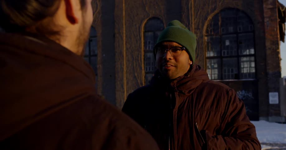 Two men chat outside with a building in the background