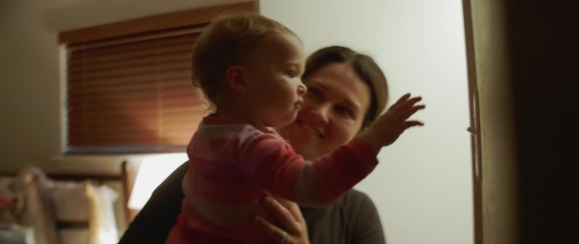 A woman holds a baby inside a house at night while the baby plays with the light switch