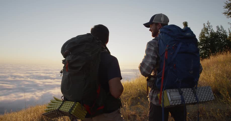 Two men with backpacks stand on a hill under a blue sky