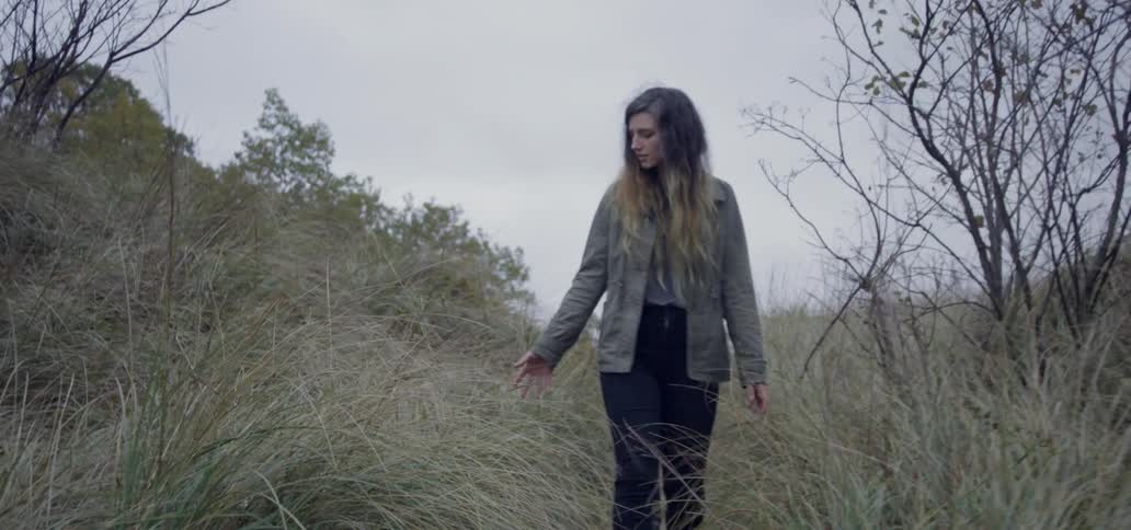 The woman is walking past the grass touching the tops
