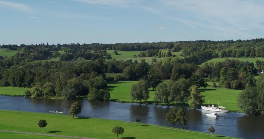 A kayak and small boat sail past a larger boat in a narrow river surrounded by green grass and trees