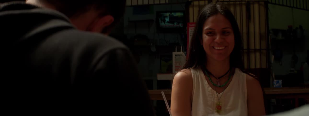 A woman sitting at a table waves and laughs at a man across the table who isn't paying attention
