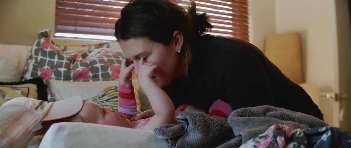 A baby is playing with its mother's eyes as she leans over it in a bedroom during the day