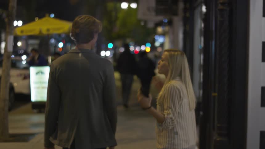 A man and woman are walking down a street at night talking