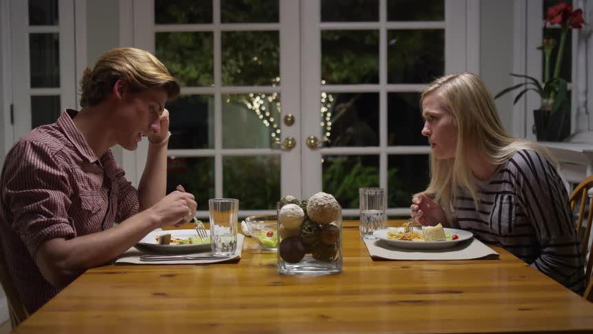 A young, blonde couple eating dinner and talking in their dining room