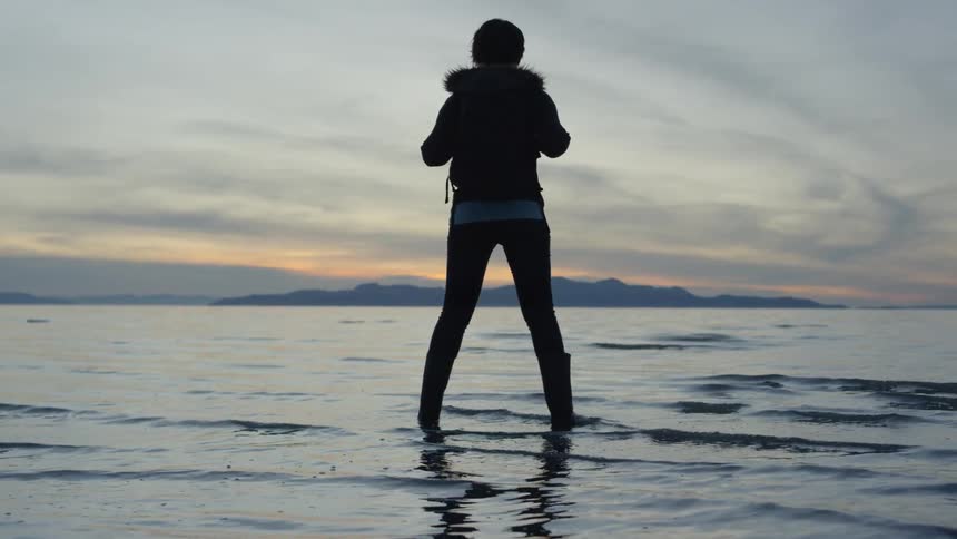 A woman stands ankle deep in the water at sunset