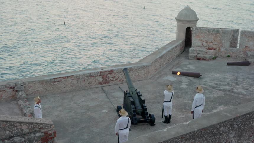 Several people stand on a fort next to water preparing to light a cannon