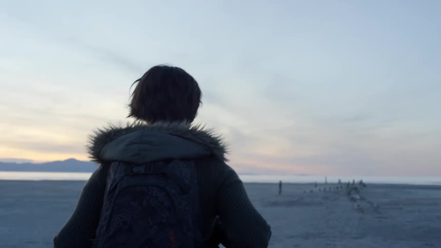 A woman wearing a backpack walks across the sand toward the water