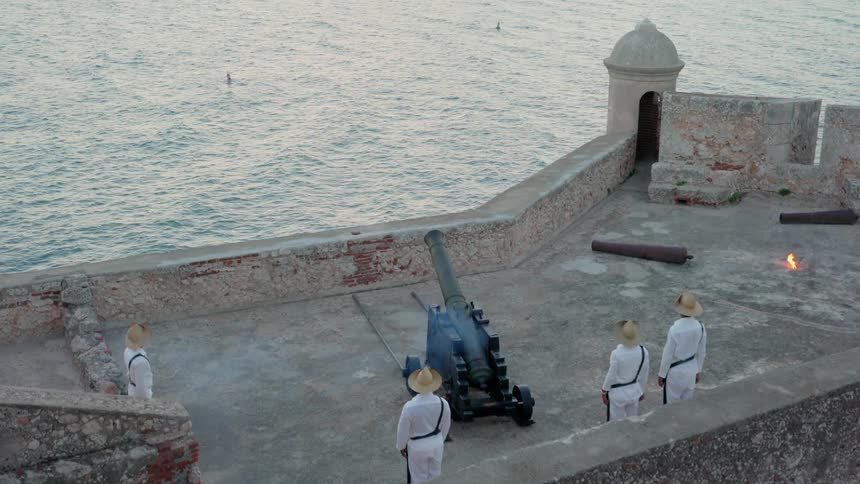 A military man lights the fuse of a cannon and it shoot over the ocean