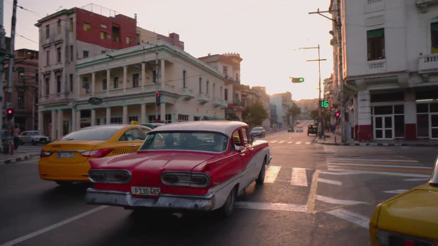 Cars drive down a busy city street with the sun shining down