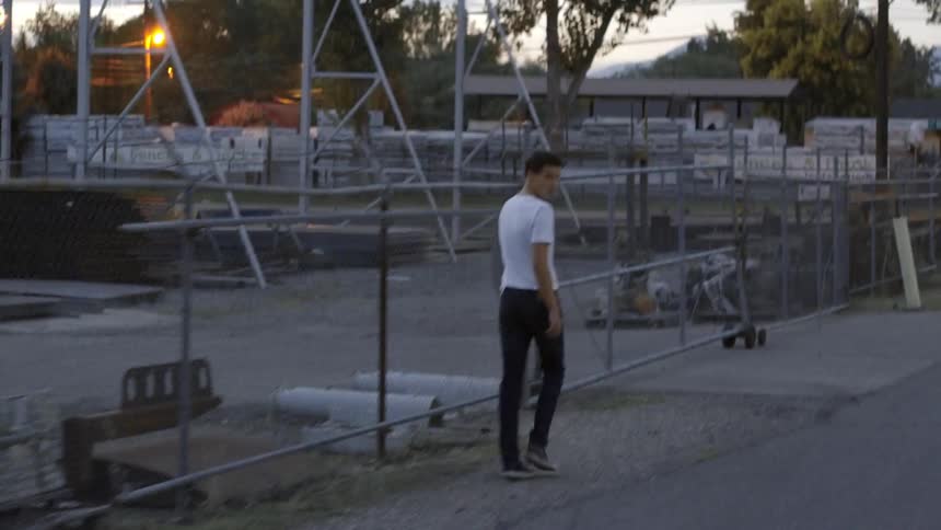 A man in a white shirt and black jeans walks down the street next to a fence