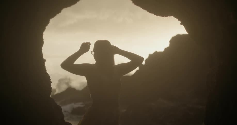 A woman standing outside a cave a the beach is lifting her arms