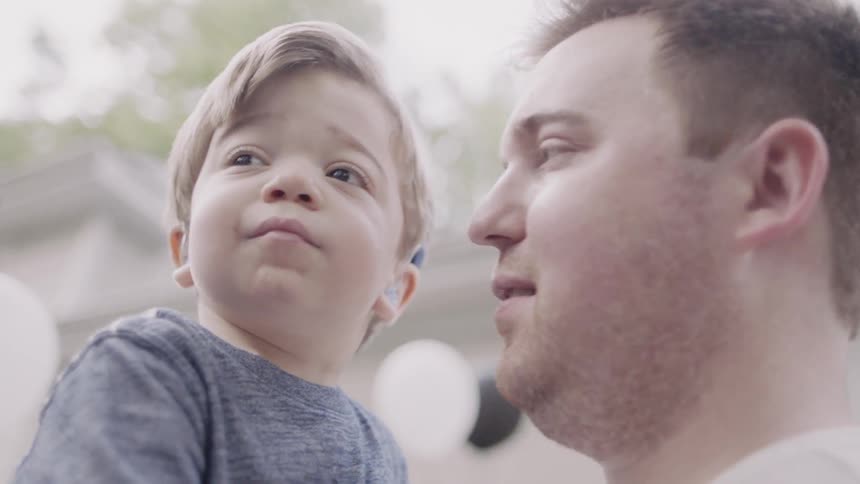 A boy wearing hearing aids is being held by his father outside a home in daylight