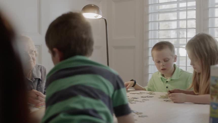 Elderly woman sits at a table with several young children as they work on a jigsaw puzzle