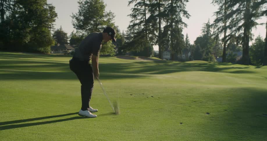 Man swings a golf club and hits a ball over the green