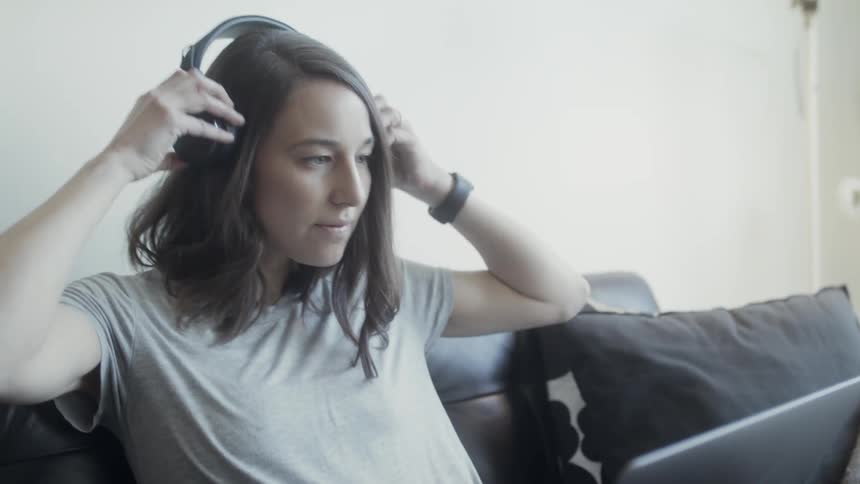 A woman is putting on headphones as she uses a laptop on a couch in daylight