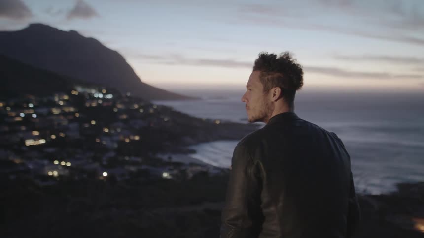 Man stands on a hilltop overlooking a city lit up below in the mountains