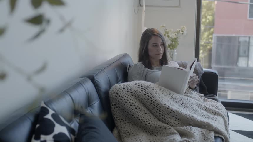 Woman sits on a couch covered in a blanket reading a book