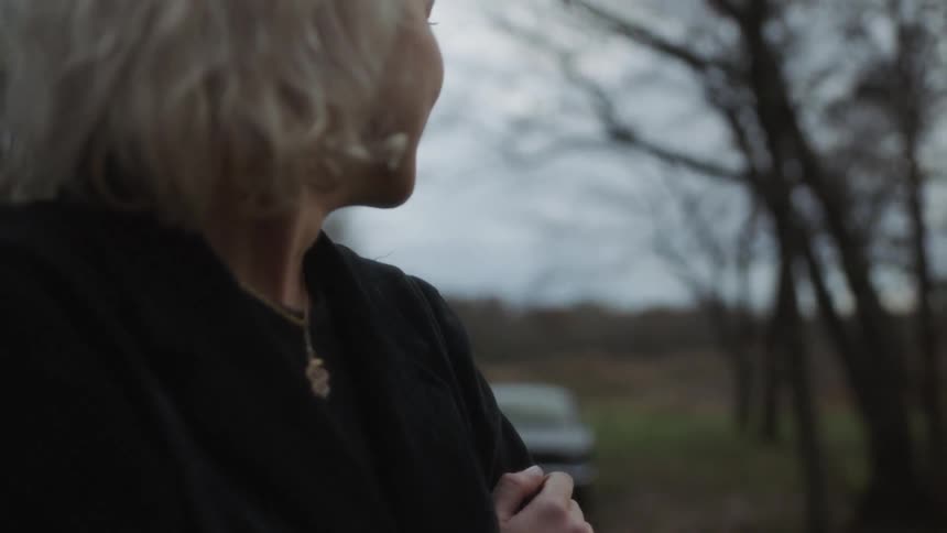 A woman is standing outside in a rural setting smiling and looking around near dusk