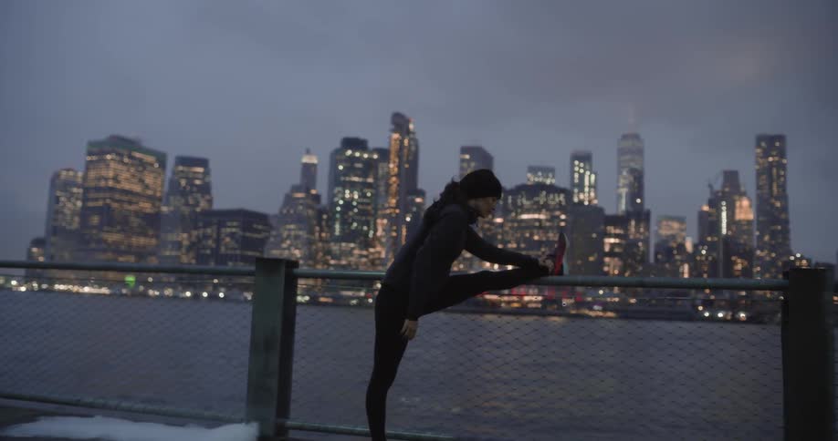 Woman is stretching her legs on fence in front of city skyline at night