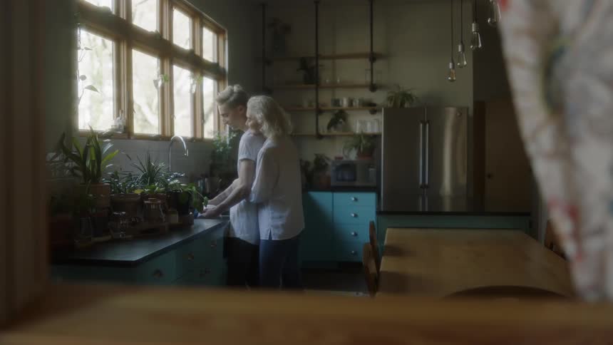 A woman hugs a man while he washes dishes