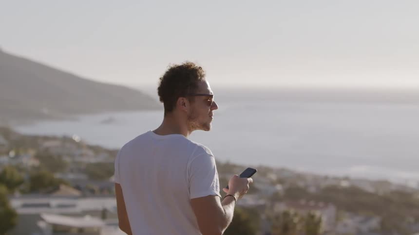A man is using his cellphone while taking in a coastal view on a sunny day
