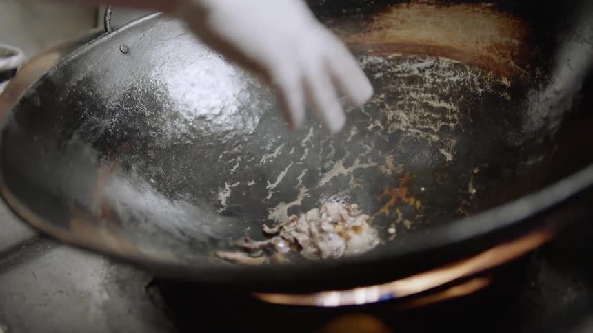 A man stands in a kitchen adding food and seasonings to a large wok