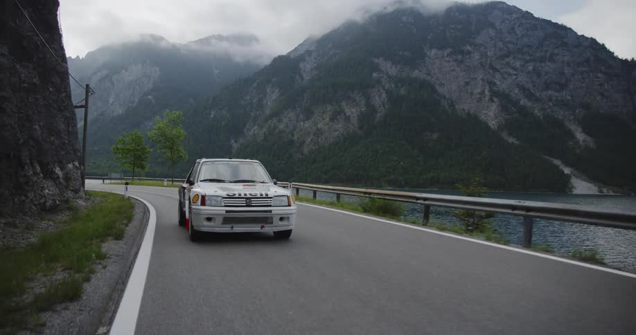 White vehicle speeds down a winding country road near a lake and mountains