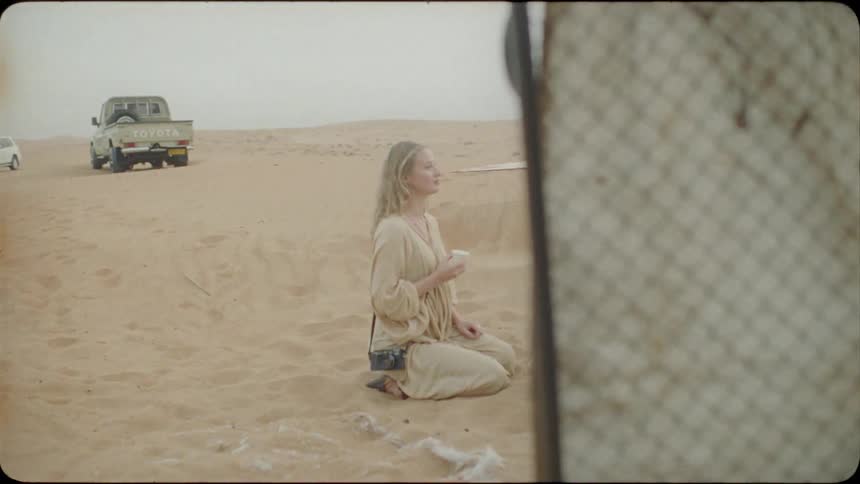 A woman and two men kneel on the sand to share a meal in the desert