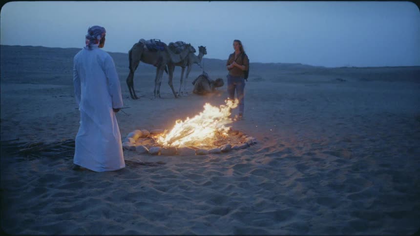 A man and woman stand near a campfire with camels waiting nearby