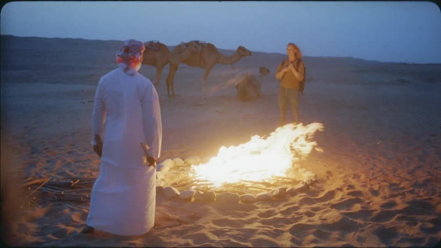 Man and woman stand by a campfire in the desert at sunset