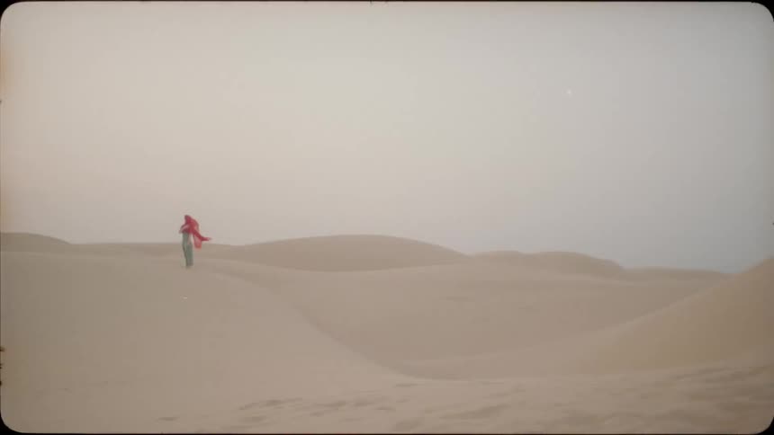 A woman is standing amid sand dunes with a scarf covering her face on a breezy day