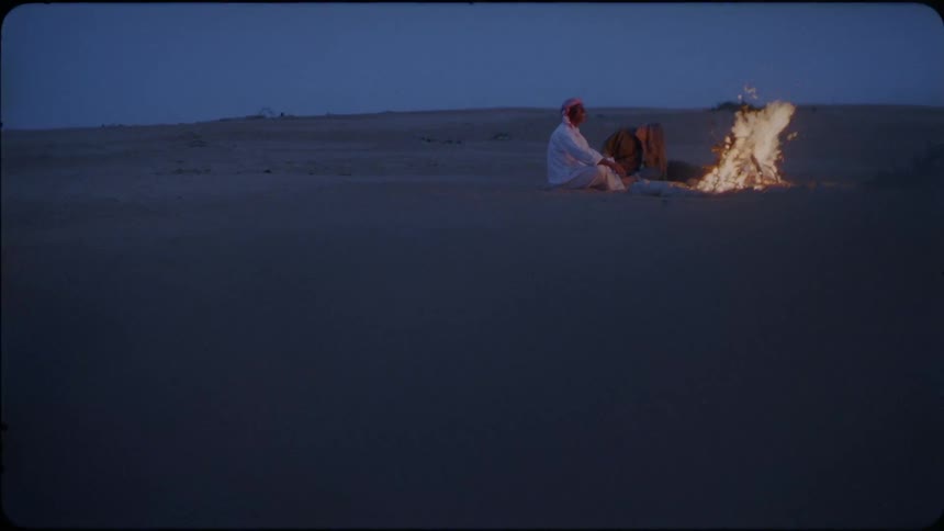 Two people sit in the desert in the evening by a campfire