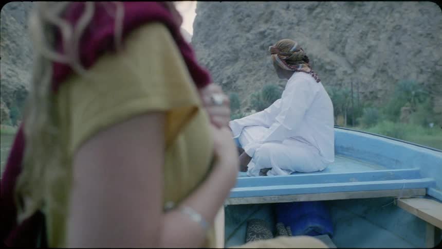 Woman stands and looks towards a man sitting on the roof of a vehicle by a lake