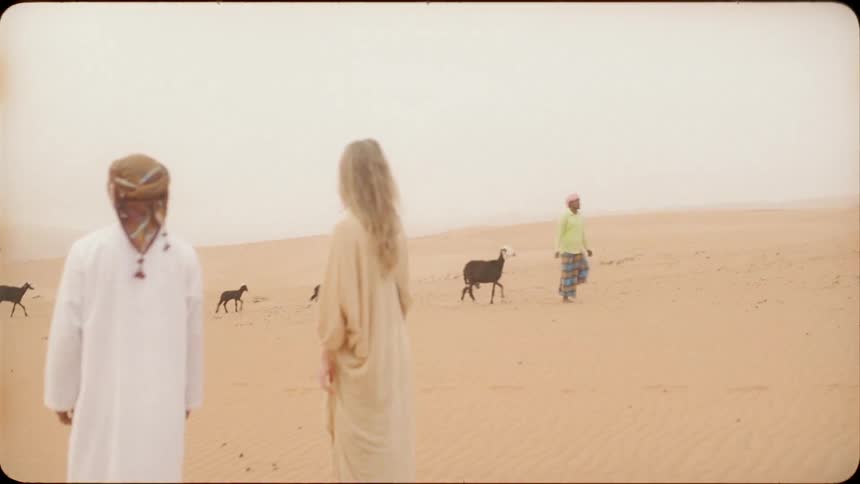 A man and woman watch as a person leads a herd of goats through the sand