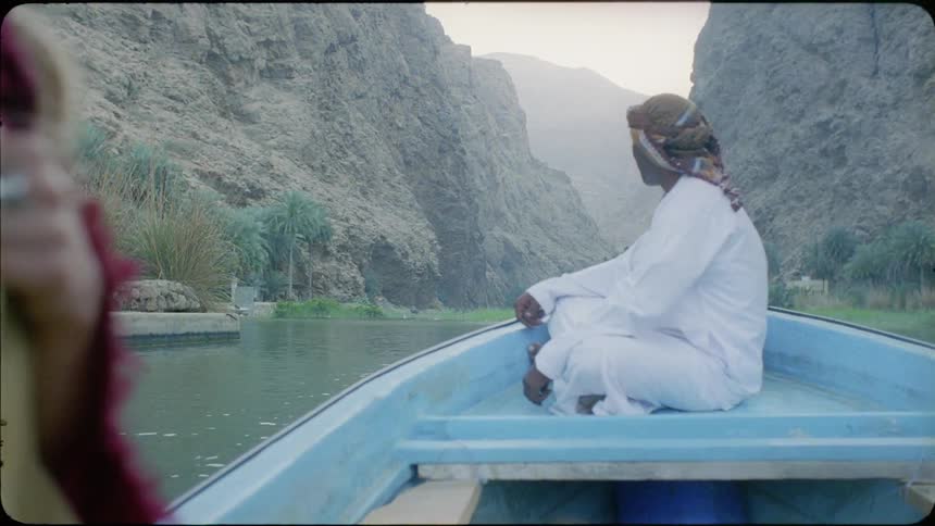Man in a white gown sits on the front of a boat as it travels down the river