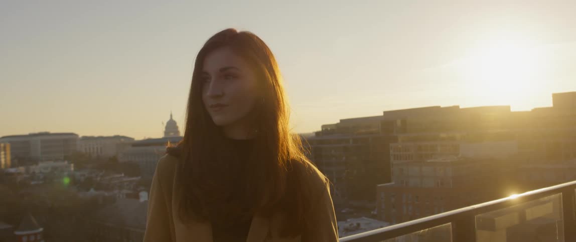 A woman is standing on a rooftop overlooking a city at sunset