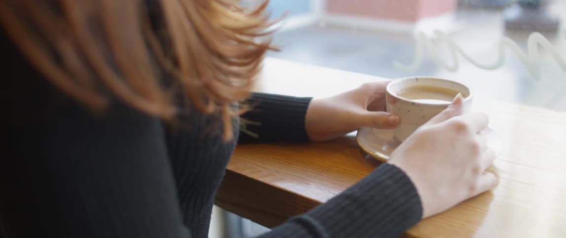 A woman sits at a coffee shop counter holding a cup of coffee