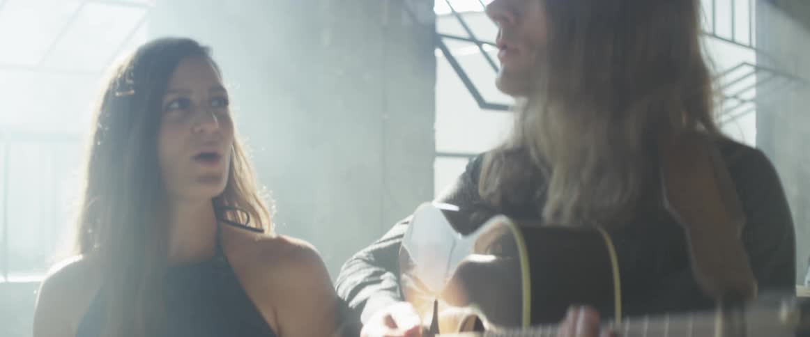 Man plays a guitar and sings with a woman while a drummer behind them drums