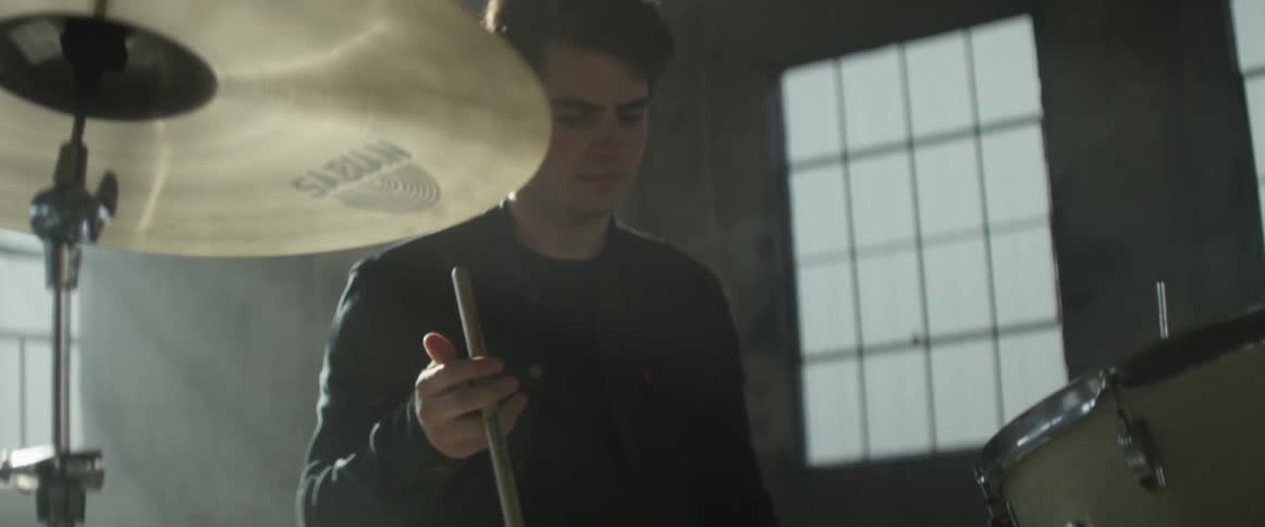 A man is holding drumsticks as he sits in a warehouse in front of a drum set