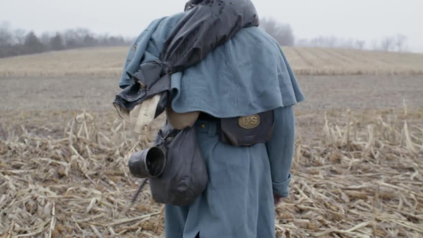 Man in a long coat walks through a field carrying a rifle
