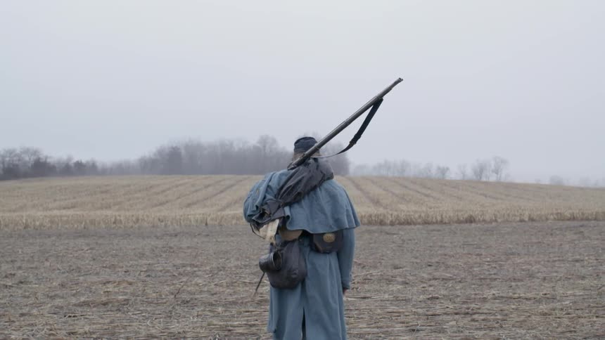 A civil war era soldier walks into a farm field carrying his rifle