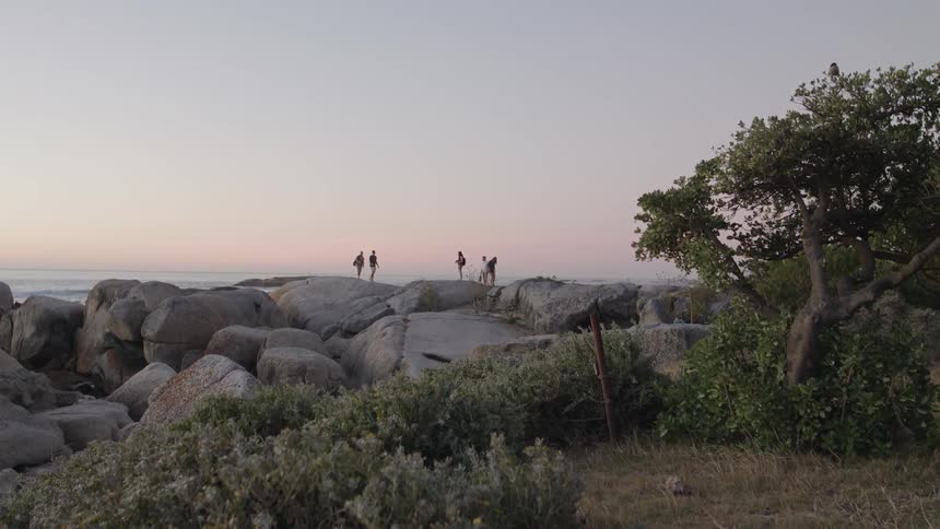 A group of people are standing on the large rocks by the shore