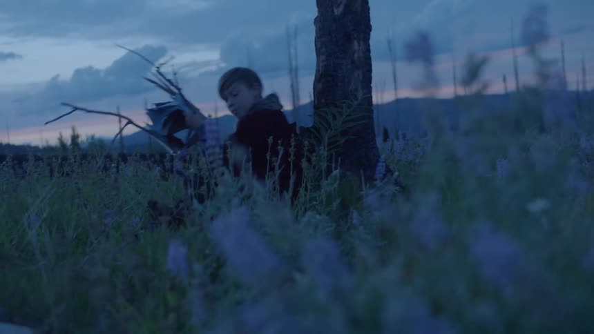 Young boy in a red cape sits down in front of a tree