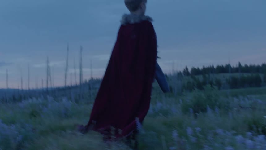 A boy wearing a cloak is holding a mask and walking in a natural setting at dusk