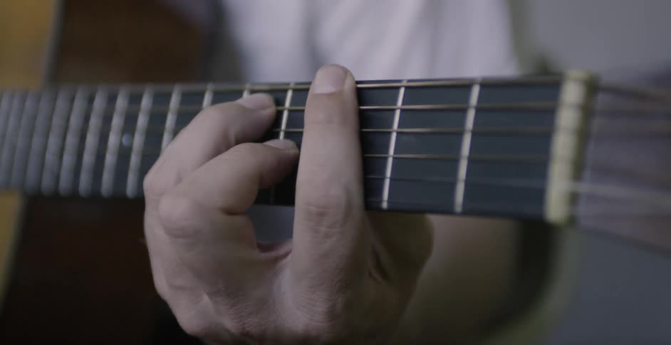 A man's fingers are pressing the strings on a guitar as he plays indoors in daylight
